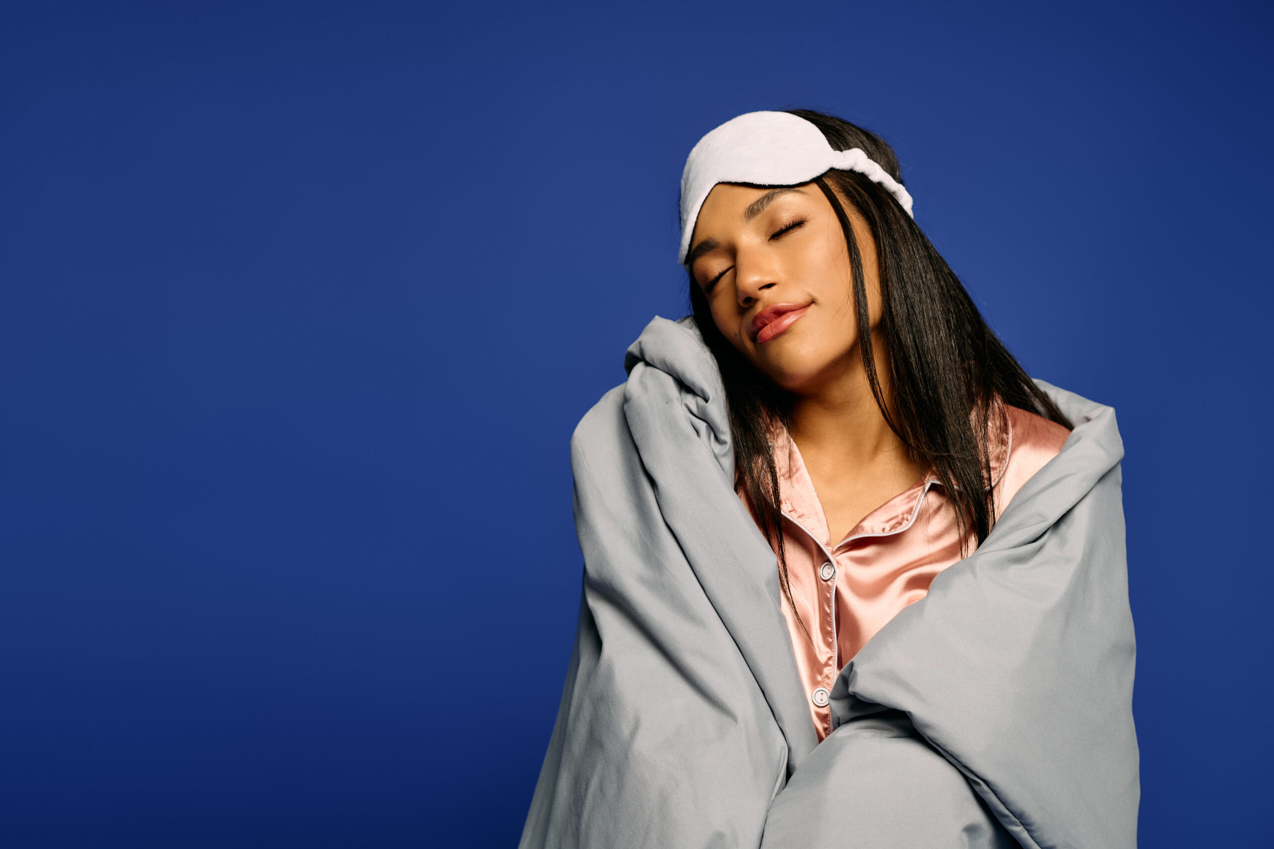 Brunette woman dressed in silk pajamas relaxes peacefully while wrapped in a cozy blanket. mulher sentada se cobrindo com lençol cinza de olhos fechados