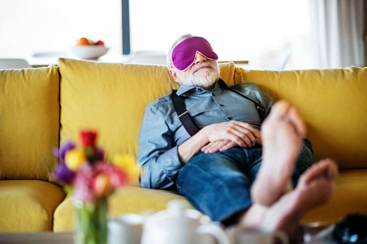 Senior man with eye mask lying on sofa indoors at home, relaxing. Senhor dormindo sentado no sofa com as pernas levantadas