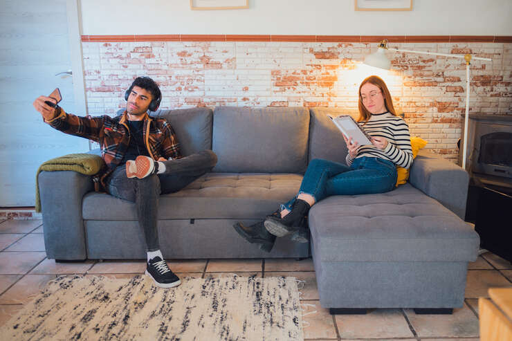 Jovens relaxando em sofá, um usando smartphone e a outra lendo.